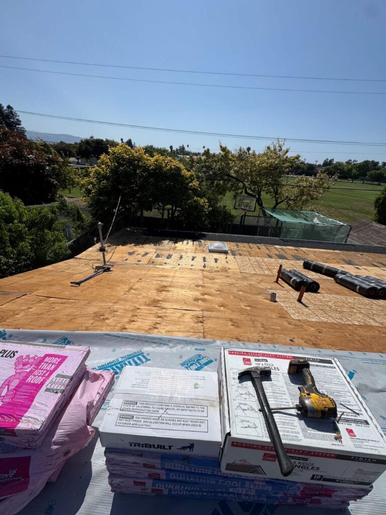 Roof replacement in progress with plywood decking exposed, synthetic underlayment, shingles, and roofing tools staged for installation.